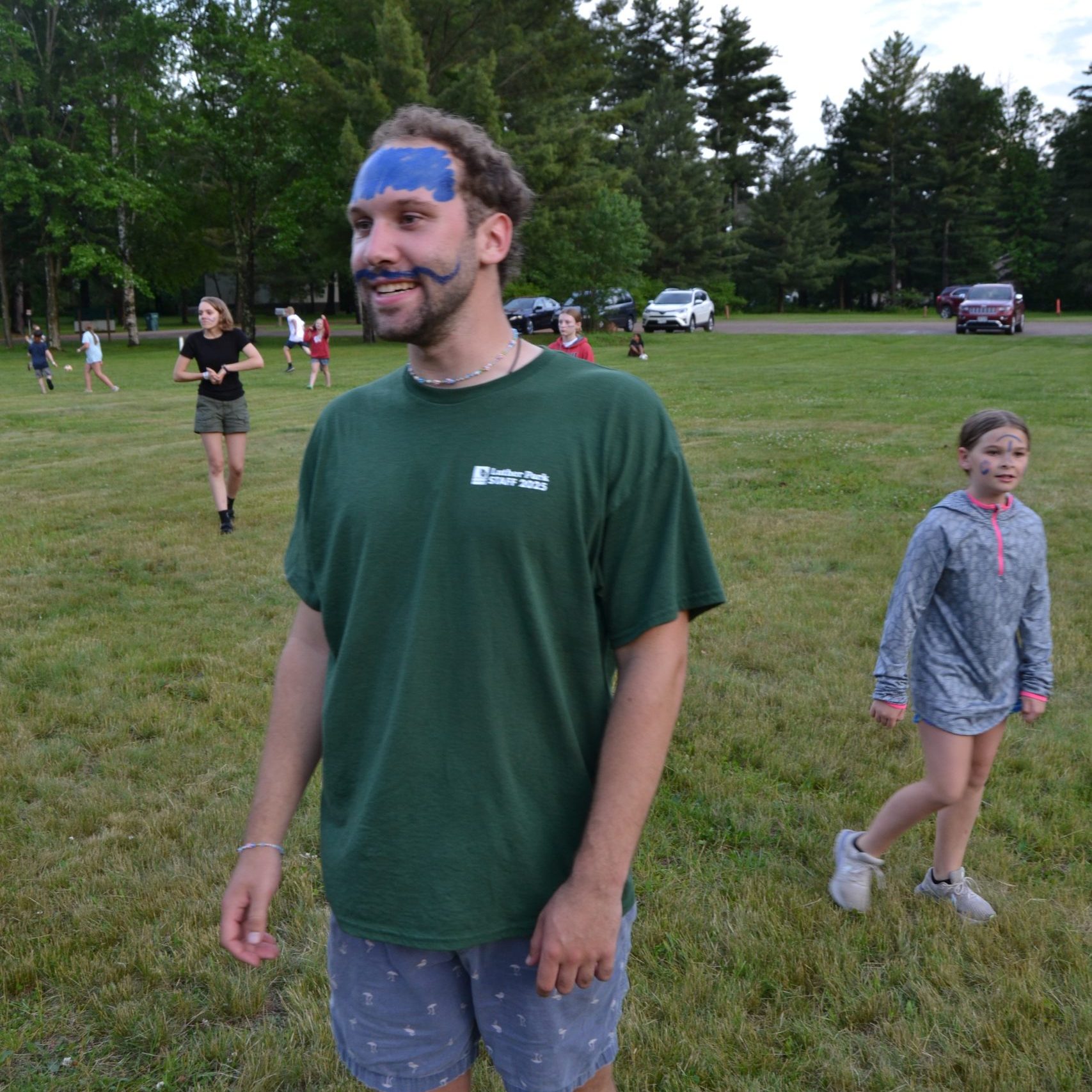 Man with blue face paint and green shirt walking in a grassy park, others playing in background.