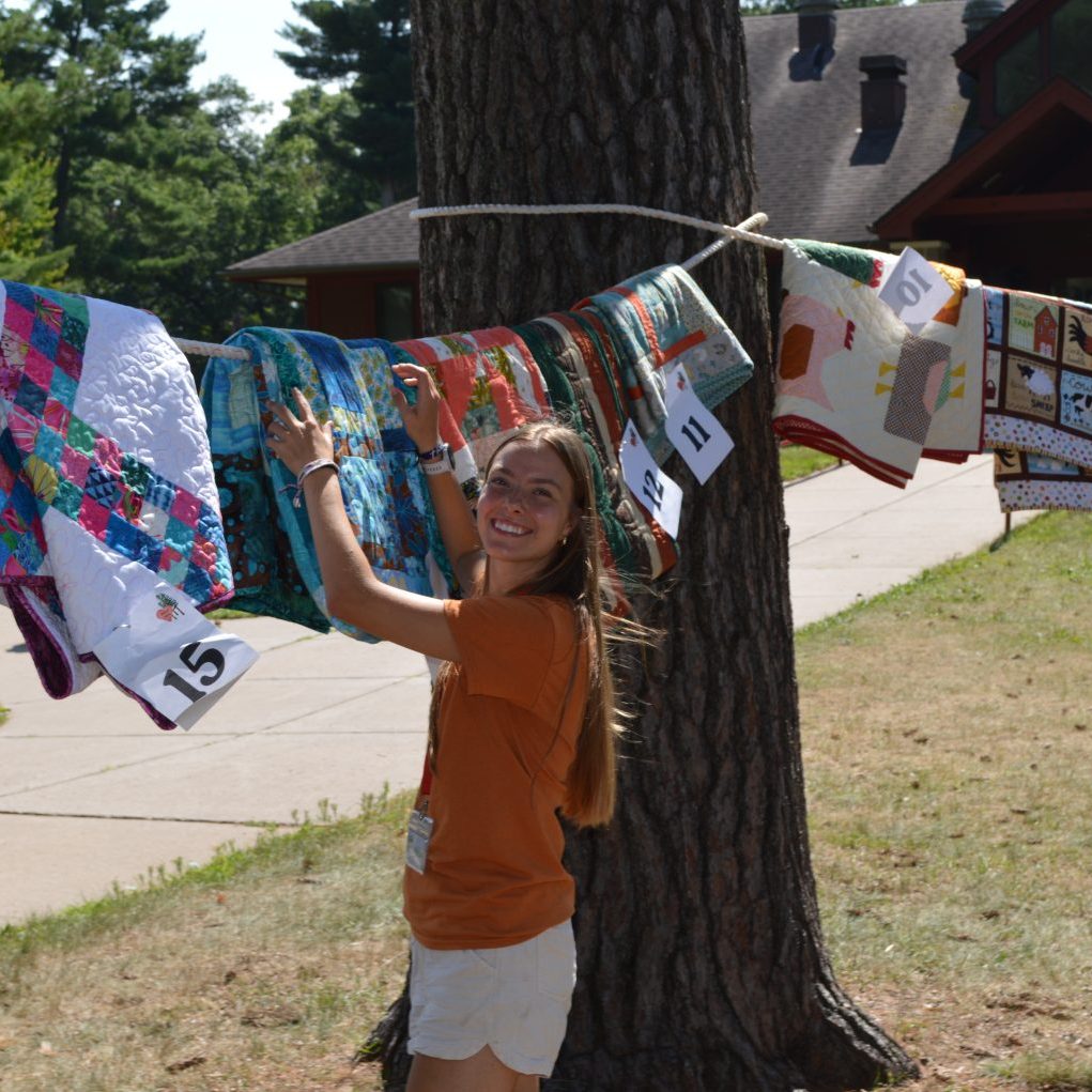 Young girl in an orange shirt holds and arranges colorful quilted blankets on a clothesline tied around a tree outdoors.