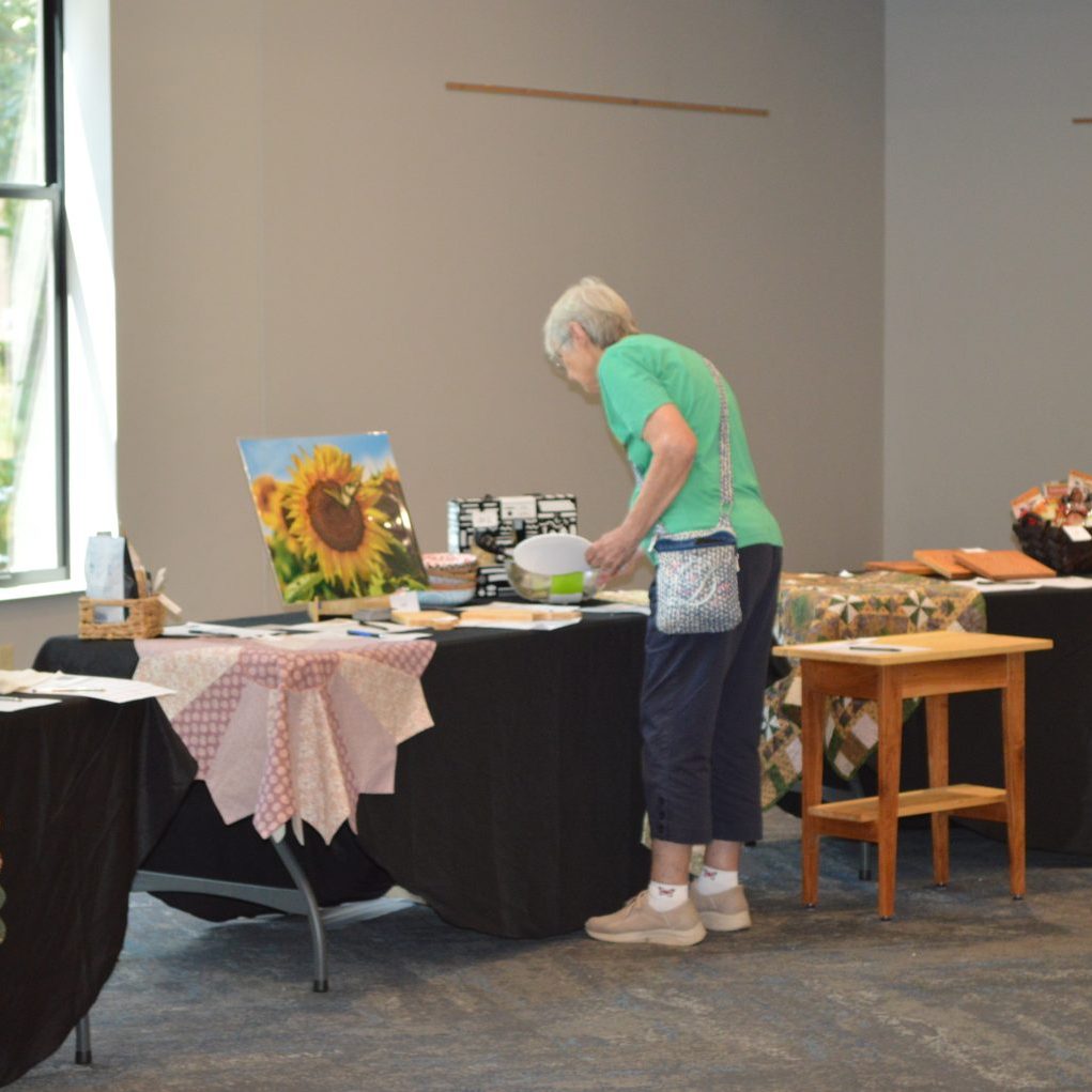 Older woman in a green shirt browsing handmade crafts on display tables, with quilts and a sunflower painting nearby.
