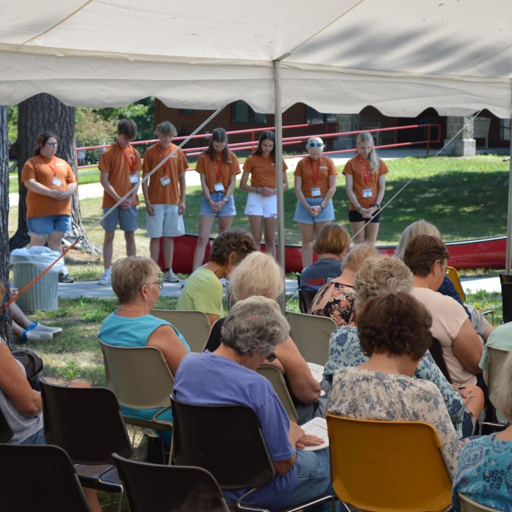 Group of young volunteers in orange shirts with name badges stand on a low stage under a white tent at an outdoor community event, facing seated audience.