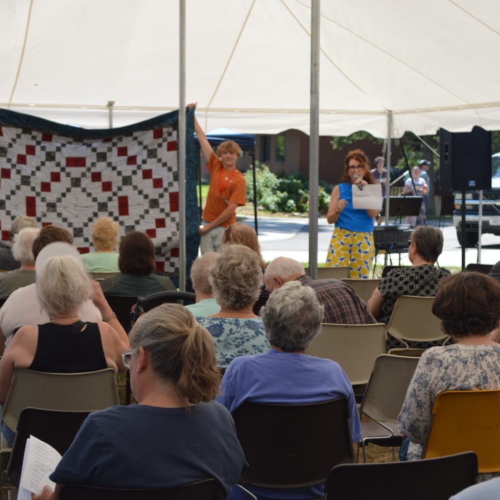 Outdoor community event under a white canopy; two volunteers hold a patchwork quilt as a speaker addresses the audience.